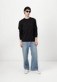 Felpa nera con polsini e orlo a costine, abbinata a jeans a gamba ampia blu chiaro e sneakers bianche. Modella con occhiali da sole neri.