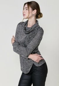 Graues Oberteil mit Kapuze und strukturierter Optik, langen Ärmeln und abgerundetem Saum, kombiniert mit schwarzen, figurbetonten Hosen. Glatter, weicher Stoff.
