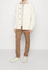 Jasno kremowa kurtka na guziki nałożona na szarą koszulę z kołnierzykiem, zestawiona z brązowymi spodniami i kremowobiałymi trampkami. Casualowa, warstwowa stylizacja.