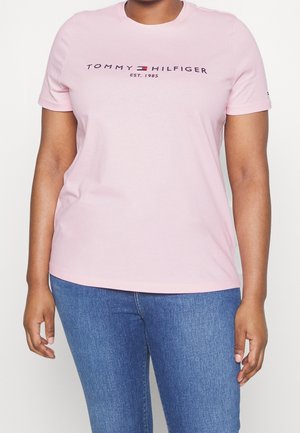 Kobieta ubrana w jasnoróżowy t-shirt Tommy Hilfiger z logo i napisem "EST. 1985," w połączeniu z niebieskimi jeansami, stojąca na białym tle.
