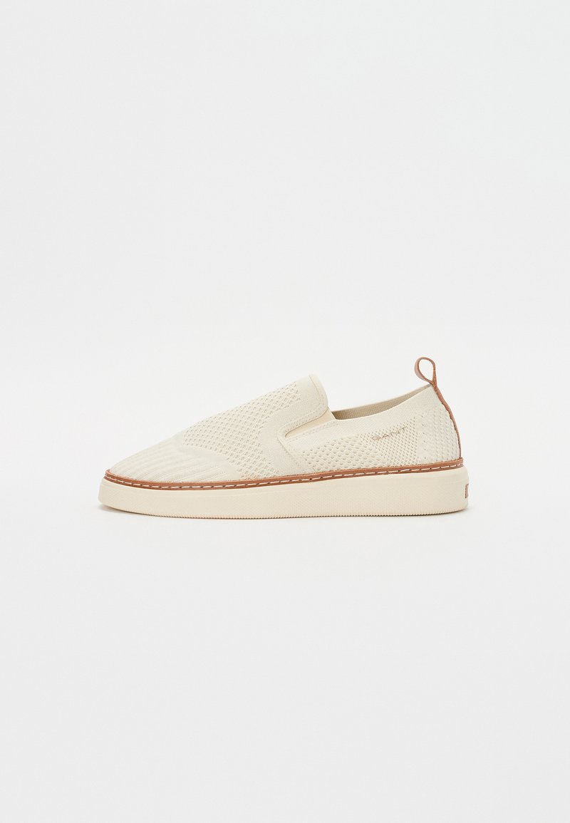 Chaussure slip-on beige avec un dessus texturé, des panneaux latéraux élastiques et un contour contrastant cousu. Dispose d'une petite languette au talon pour un enfilage facile.