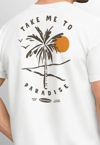 Weißes T-Shirt mit Grafik einer Palme, Sonnenuntergang in Orange und dem Text "NIMM MICH MIT INS PARADIES" in Braun. Zusätzliche Logos unten.