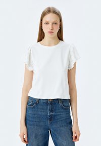Witte T-shirt met korte mouwen en broderie anglaise details op de mouwen, gecombineerd met hoge taille denim jeans. Glad katoenen textuur.
