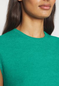 Femme aux longs cheveux bruns portant un pull sans manches côtelé vert, vue du cou à la poitrine sur un fond uni.