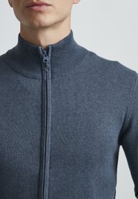 Mørkeblå, glidelåsgenser med høy ribbestrikket krage, glatt tekstur og tettsittende design. Sømmer er synlige langs skuldrene.