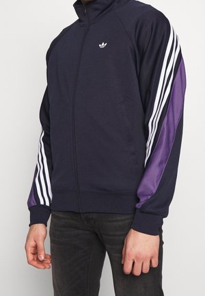 Man draagt een zwart Adidas zip-up jack met witte strepen en paarse panelen op de mouwen, gecombineerd met donkere spijkerbroek.