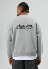 Grijze trui met ronde hals, met zwarte tekst op de achterkant: "RAÍCES VIVAS" en afbeeldingen. Gestructureerde stof met geribbelde manchetten.