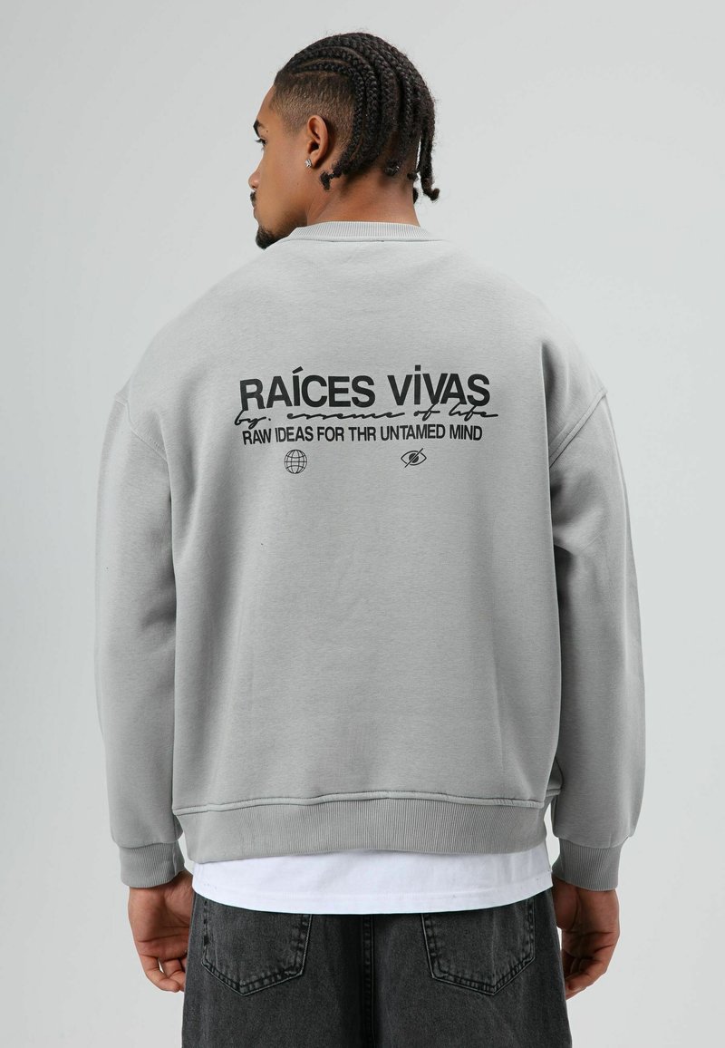 Grijze trui met ronde hals, met zwarte tekst op de achterkant: "RAÍCES VIVAS" en afbeeldingen. Gestructureerde stof met geribbelde manchetten.