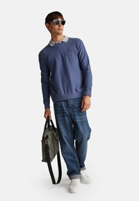 Niebieski sweter dzianinowy z szarym kołnierzem, luźne niebieskie dżinsy, białe trampki i szara torba z czarnymi paskami; noszone ciemne okulary.