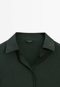 Chemise vert foncé à col avec un tissu texturé, fermeture à boutons sur le devant, et une étiquette lisant "Massimo Dutti Made in China" à l'intérieur du col.