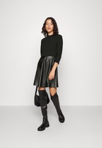 Pull en maille texturée vert foncé à manches trois-quarts, associé à une jupe évasée en simili cuir noir et des bottes noires jusqu'aux genoux.