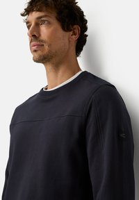 Sweat-shirt bleu marine en tissu doux et texturé, doté d'un col rond, d'une couture horizontale et d'un patch logo sur la manche gauche.