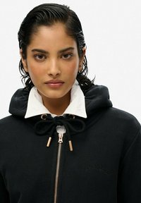 Felpa nera con collo bianco, caratterizzata da coulisse e dettagli in oro. Il logo del marchio è impresso sul lato sinistro.