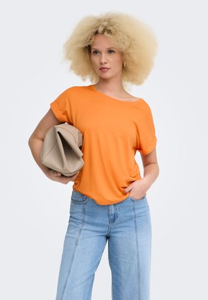 Femme aux cheveux blonds bouclés portant un haut orange à manches courtes et un jean bleu clair, tenant une pochette beige à rabat sous un bras.