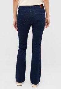 Granatowe dzwony z gładką teksturą, dwoma tylnymi kieszeniami i brązową skórzaną metką na pasie. Wykonane z dżinsowej tkaniny.