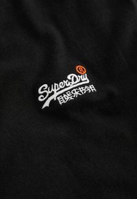 Țesătură neagră cu logo brodat alb și portocaliu, cu o textură netedă. Designul include text stilizat și caractere asiatice.