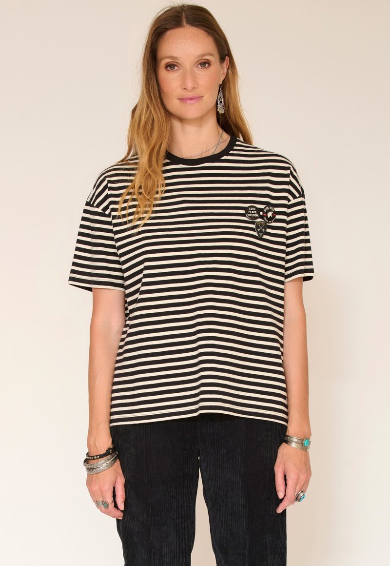 T-shirt rayé noir et blanc avec un col rond, des manches courtes, et des patchs brodés en forme de cœur et de bouclier sur la poitrine.
