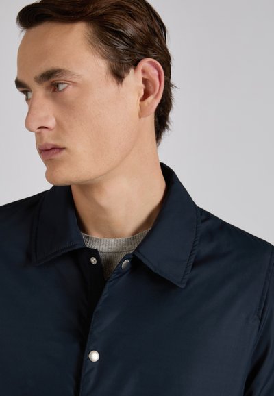 Joven con cabello castaño corto, vistiendo una chaqueta azul marino con botones y un suéter de punto gris, mirando hacia la izquierda contra un fondo liso.