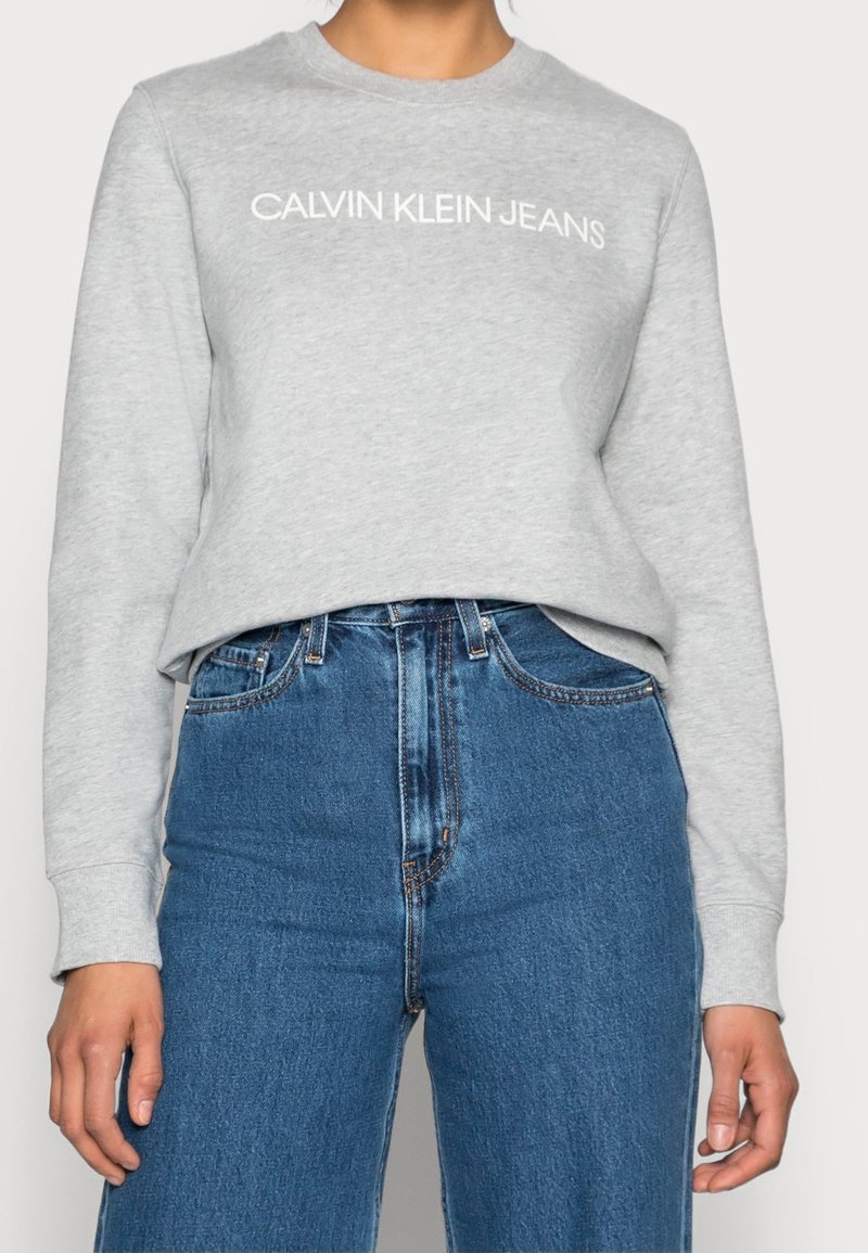 Personne portant un sweat-shirt Calvin Klein Jeans gris clair et un jean bleu taille haute, debout les bras détendus le long du corps.