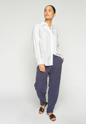 Nő fehér hosszú ujjú inget, bő lila nadrágot és sötétbarna ujjnyi nyitott szandált visel, és egy egyszerű világos háttér előtt áll.