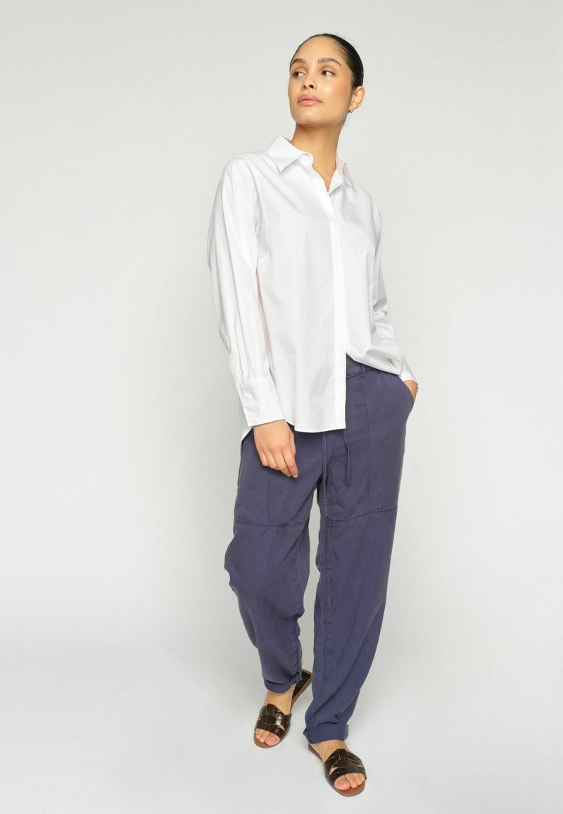 Naine, kes kannab valget pika varrukaga särki, lahtiseid lillaid pükse ja tumedat pruunide varbavahe sandaale, seisab tavalise hele tausta ees.