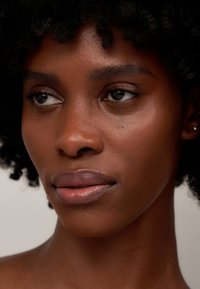 Primer plano de un rostro con piel oscura, cabello negro rizado, tez suave, color de labios natural y ojos claros; sin maquillaje ni adornos visibles.