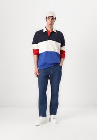 Tovefarget pullover med marineblå, kremfargede og røde striper, sammen med blå jeans og hvite sneakers; en lyse farget caps fullfører looket.