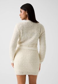 Robe-pull en maille crème avec un motif texturé. Caractéristiques : manches longues, silhouette ajustée et ceinture côtelée à la taille.
