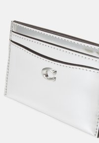 Štíhlý stříbrný držák karet se třemi p řihrádkami na karty a stříbrným metalickým logem "C" na přední kapse.