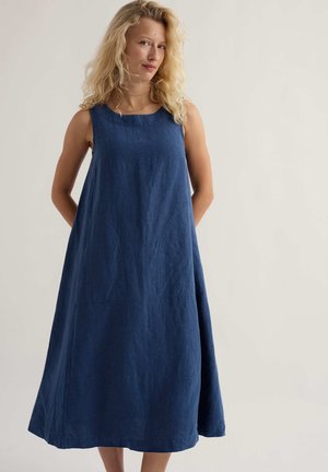 Szőke hajú nő, aki ujjatlan, laza, térdig érő kék ruhát visel, és egyszerű, világos háttér előtt áll.