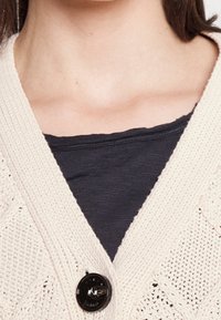 Cárdigan de punto beige con escote en V, que presenta un detalle de botón negro. Debajo, se puede ver una camisa azul marino texturizada.