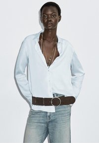 Camicia button-up azzurro chiaro con maniche lunghe, abbinata a denim chiaro. Cintura in pelle scamosciata marrone con una grande fibbia rotonda argentata. Diverse collane a strati.