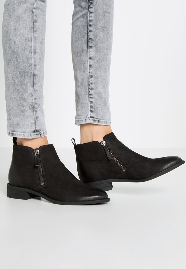 Bottines noires en suédine synthétique avec bouts pointus, fermetures éclair latérales et petits talons carrés, associées à un jean gris clair ajusté.