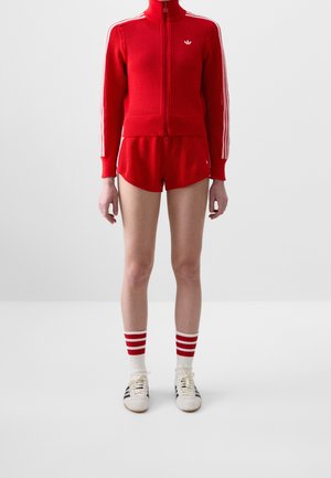 Personne portant une veste en maille rouge zippée et un short assorti, des chaussettes blanches avec des rayures rouges, et des baskets blanches avec des rayures noires.