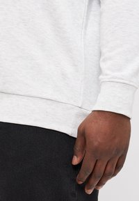 Grauer Pullover mit gerippten Bündchen und Saum, neben einem dunklen Stoff präsentiert. Die Textur wirkt weich und leicht meliert.