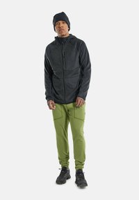 Must have kapuutsiga must fliiskrae, millele on sobitatud rohelised jogger-püksid esi-taskutega. Model kannab musti spordijalatsid ja rätikut.
