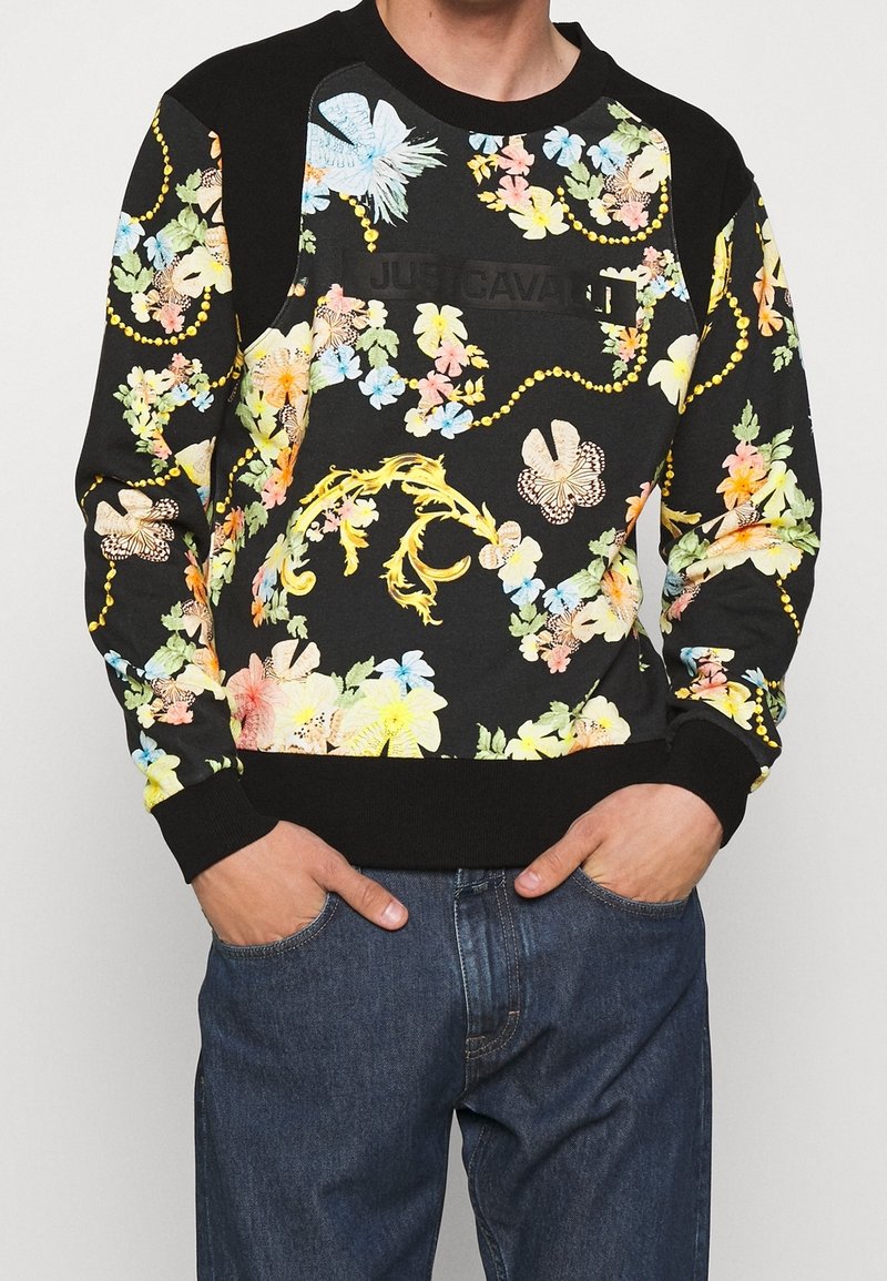 Sweat-shirt noir avec motifs floraux colorés et papillons, chaînes semblables à des perles jaunes, et logo "Just Cavalli", porté par une personne les mains dans les poches d'un jean bleu.