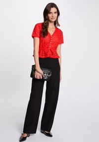 Haut au crochet rouge à manches courtes et décolleté en V, avec des boutons et un lien à la taille, associé à un pantalon large noir et un sac noir.