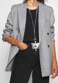 Blazer a cuadros gris con diseño de doble botonadura, combinado con una camiseta negra con relieve y jeans negros. Un mini accesorio plateado cuelga de una correa.