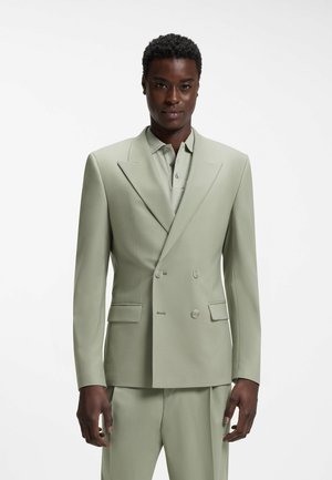 Man draagt een lichtgroene dubbelgeknoopte blazer over een overhemd met kraag en bijpassende broek, staand tegen een effen witte achtergrond.
