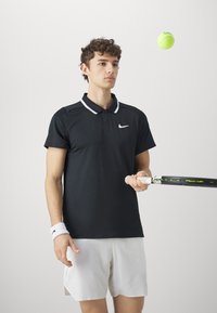 Juodas Nike polo marškinėlis su baltu apykakle, vilkimas su baltais šortais. Asmuo laiko teniso raketę, o šalia skraido žalias teniso kamuoliukas.