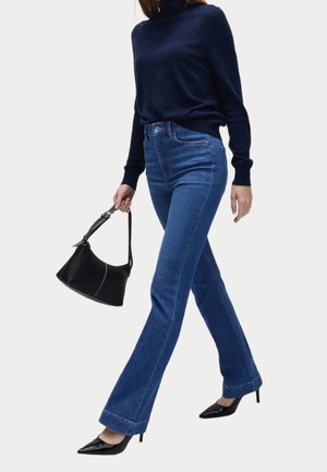 Vrouw die loopt en een marineblauwe coltrui draagt, blauwe jeans met hoge taille, zwarte puntige hakken en een zwarte handtas met een gestikte band vasthoudt.