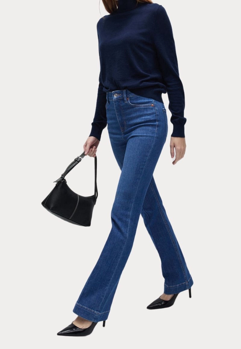Femme marchant portant un col roulé bleu marine, un jean taille haute bleu, des talons noirs pointus, et tenant un sac à main noir avec une bandoulière cousue.