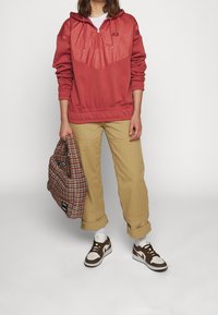 Červená mikina s texturovanou částí na hrudi, béžové kalhoty a hnědé a bílé tenisky. Drží kostkovanou tašku s šachovnicovým vzorem.