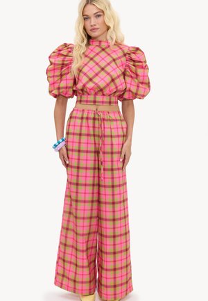 Femme portant une tenue à carreaux rose, verte et marron avec des manches courtes bouffantes et un pantalon à jambes larges, accessoirisée de bracelets colorés.