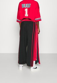 Rotes, kurzärmeliges Crop-Top mit "BUGS" und der Nummer "1" in Türkis. Schwarze, weite Hosen mit roten und weißen Seitenstreifen. Weiße Sneakers.
