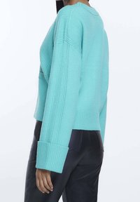 Tyrkysový pletený svetr s ribovanou texturou, spadlými rameny a širokými manžetami. Krátký, uvolněný střih v kombinaci s elegantními černými kalhotami.