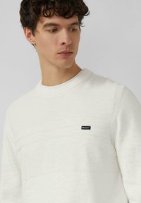 Jeune homme aux cheveux bouclés et foncés portant un pull blanc texturé à col rond avec une petite étiquette "Elevated Casual" sur la poitrine.