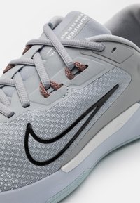 Gros plan sur une chaussure de sport Nike grise et noire avec un dessus texturé, des lacets gris clair, et du texte sur la languette et la semelle.