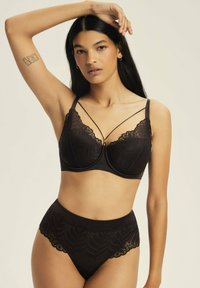 Kobieta z długimi ciemnymi włosami, nosząca czarny koronkowy stanik i pasujące majtki o wysokim stanie, podnosi jedną rękę, by dotknąć swojej głowy.